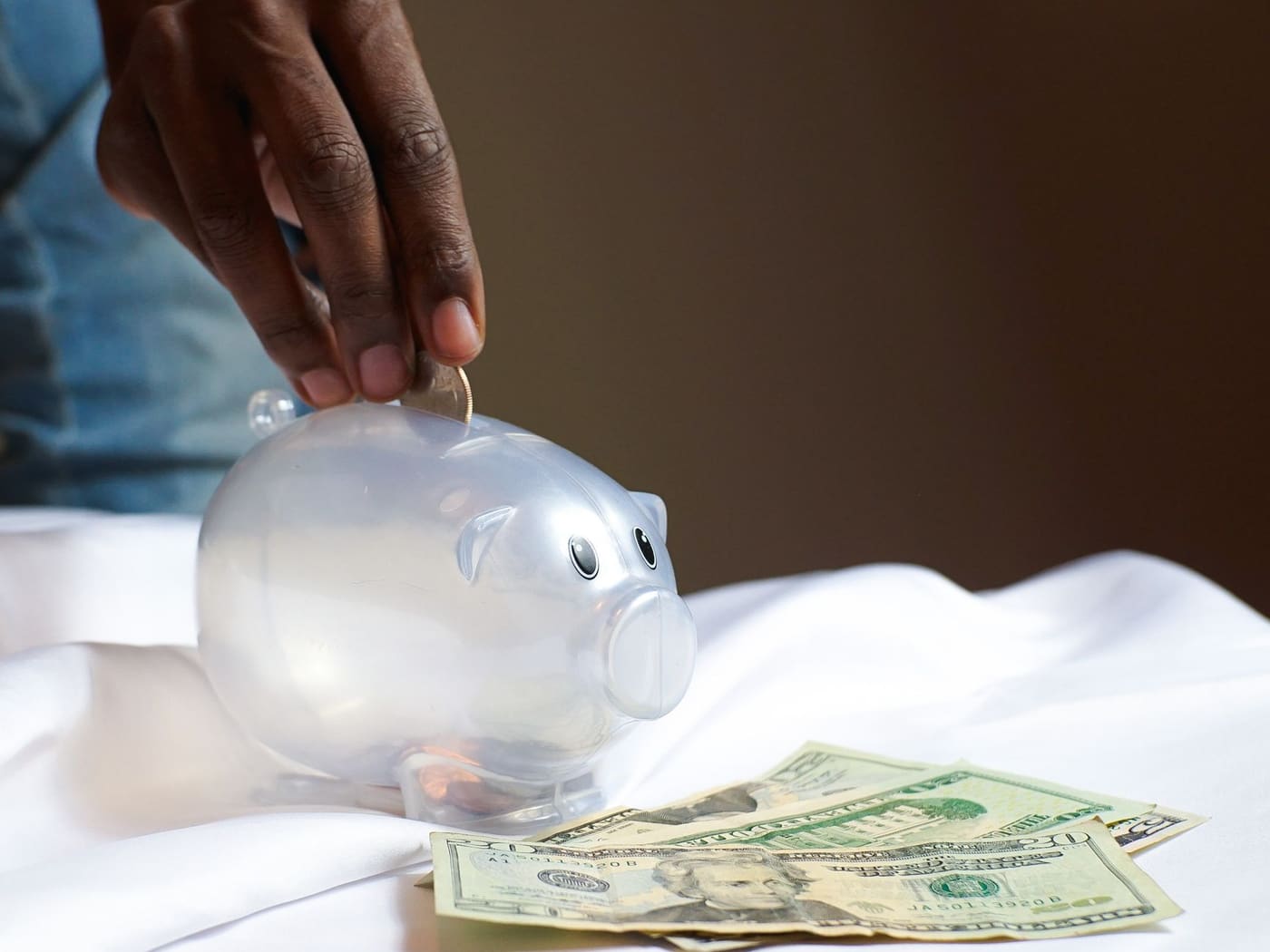 A hand placing a coin into a piggy bank beside some dollar bills